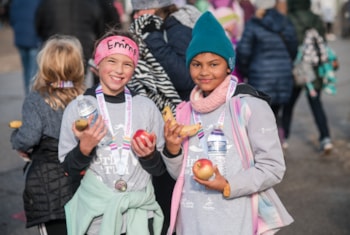 Two Girls on the Run participants smile in grey GOTR shirts holding fruit at 5K